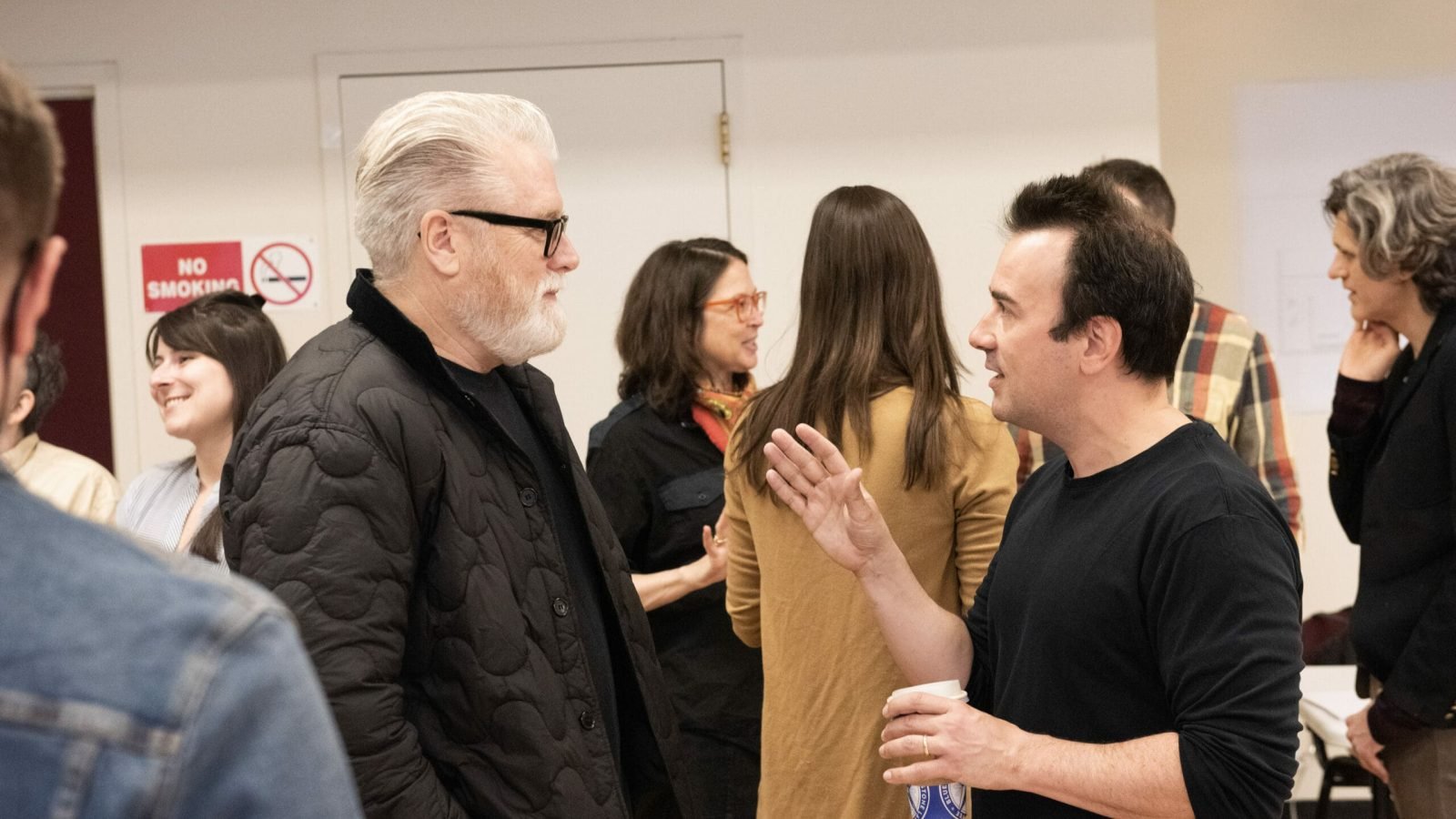 Two men converse at a meeting, other attendees in the background.