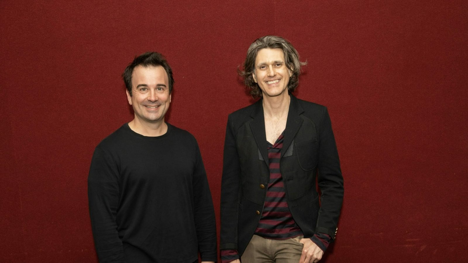 Two men smiling against a red background.