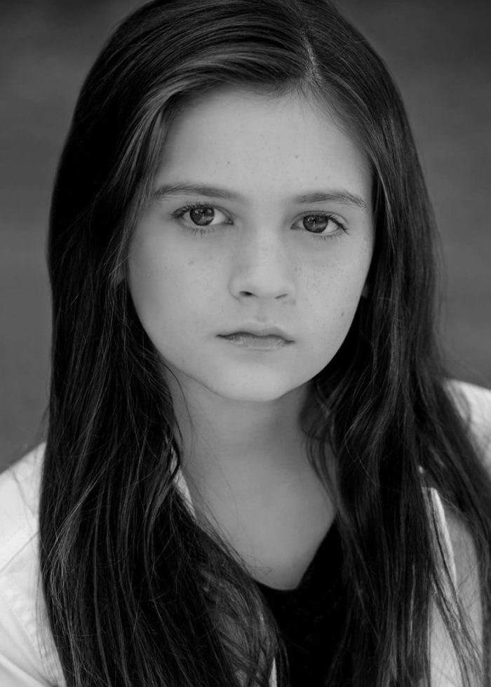 Young girl with long dark hair and freckles stares at the camera.