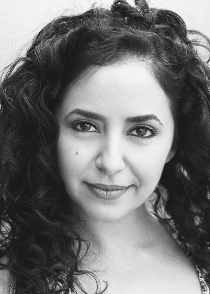 Sharone Sayegh with curly hair smiles at the camera in a black and white portrait.
