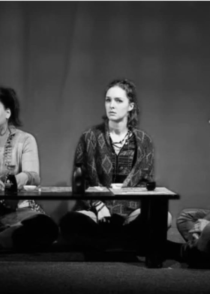 Actors in a theater production seated around a low table, reviewing 'Happy Hour'.