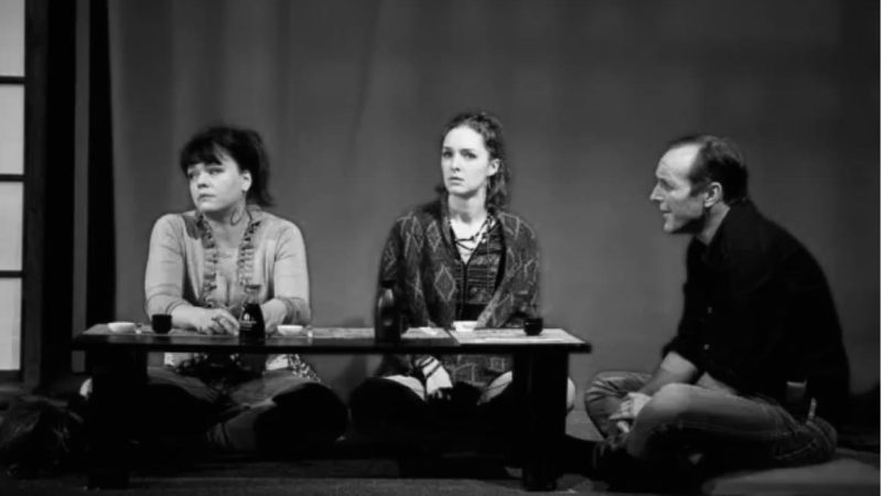Actors in a theater production seated around a low table, reviewing 'Happy Hour'.