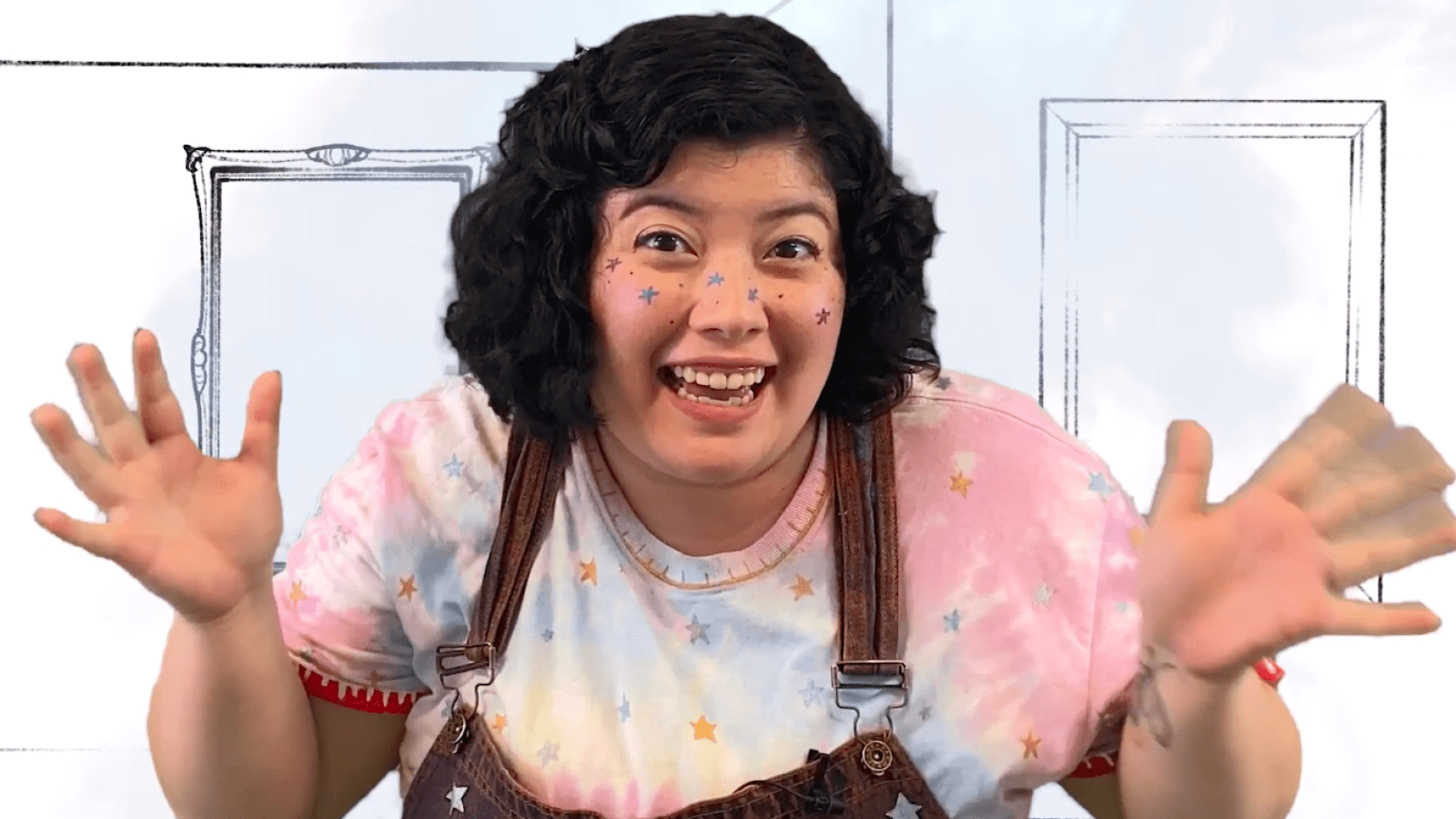 Smiling woman with star freckles, gesturing excitedly. She's wearing a tie-dye shirt and overalls.