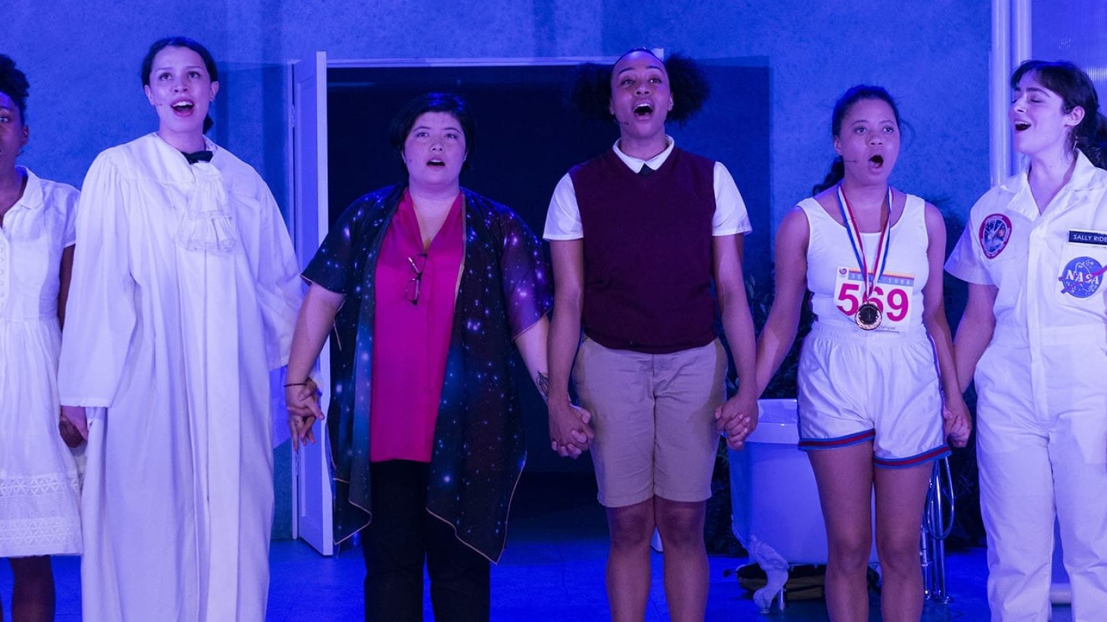 Six young women in various costumes hold hands and sing in a blue-lit room. "She Persisted" musical.