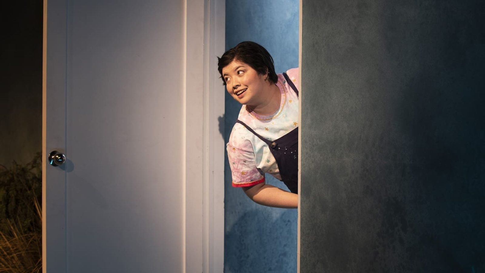A woman peeks around a corner, smiling mischievously. She's wearing a pastel top and overalls.
