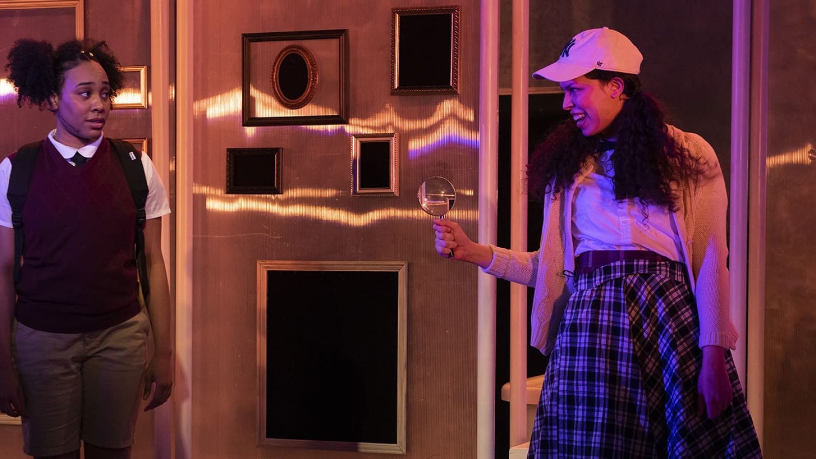 Two young women in school uniforms, one holding a magnifying glass, in a theatrical scene from *She Persisted, The Musical*.