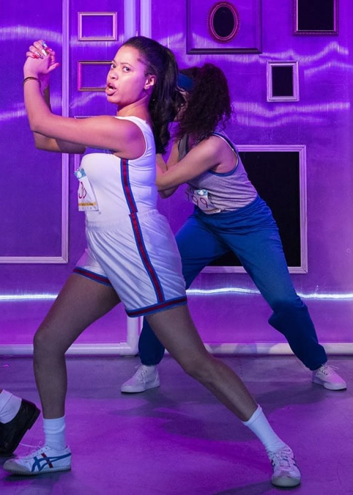 Cast of "She Persisted, The Musical" performing a dance routine, with purple lighting and a stage set.