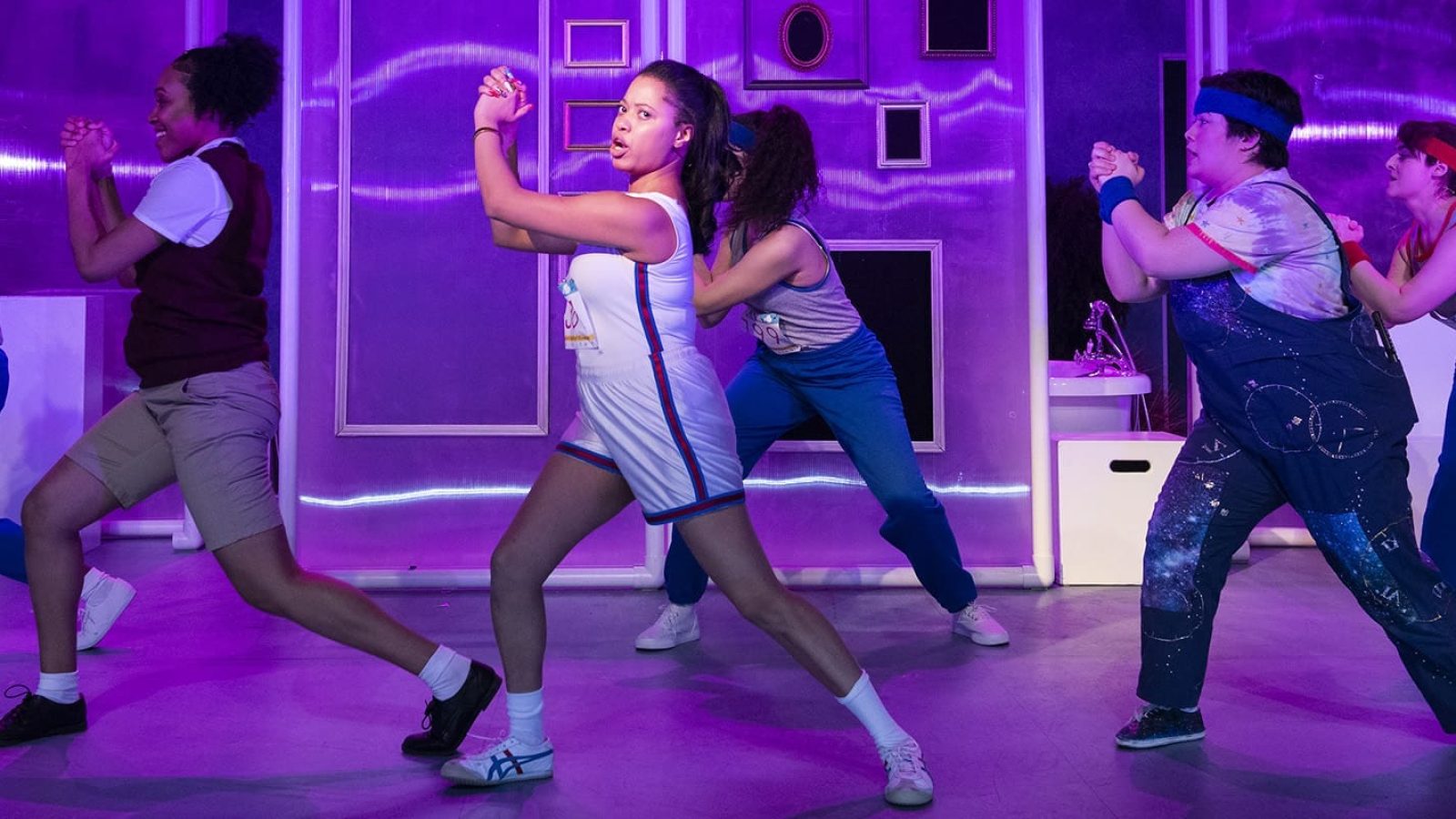 Cast of "She Persisted, The Musical" performing a dance routine, with purple lighting and a stage set.