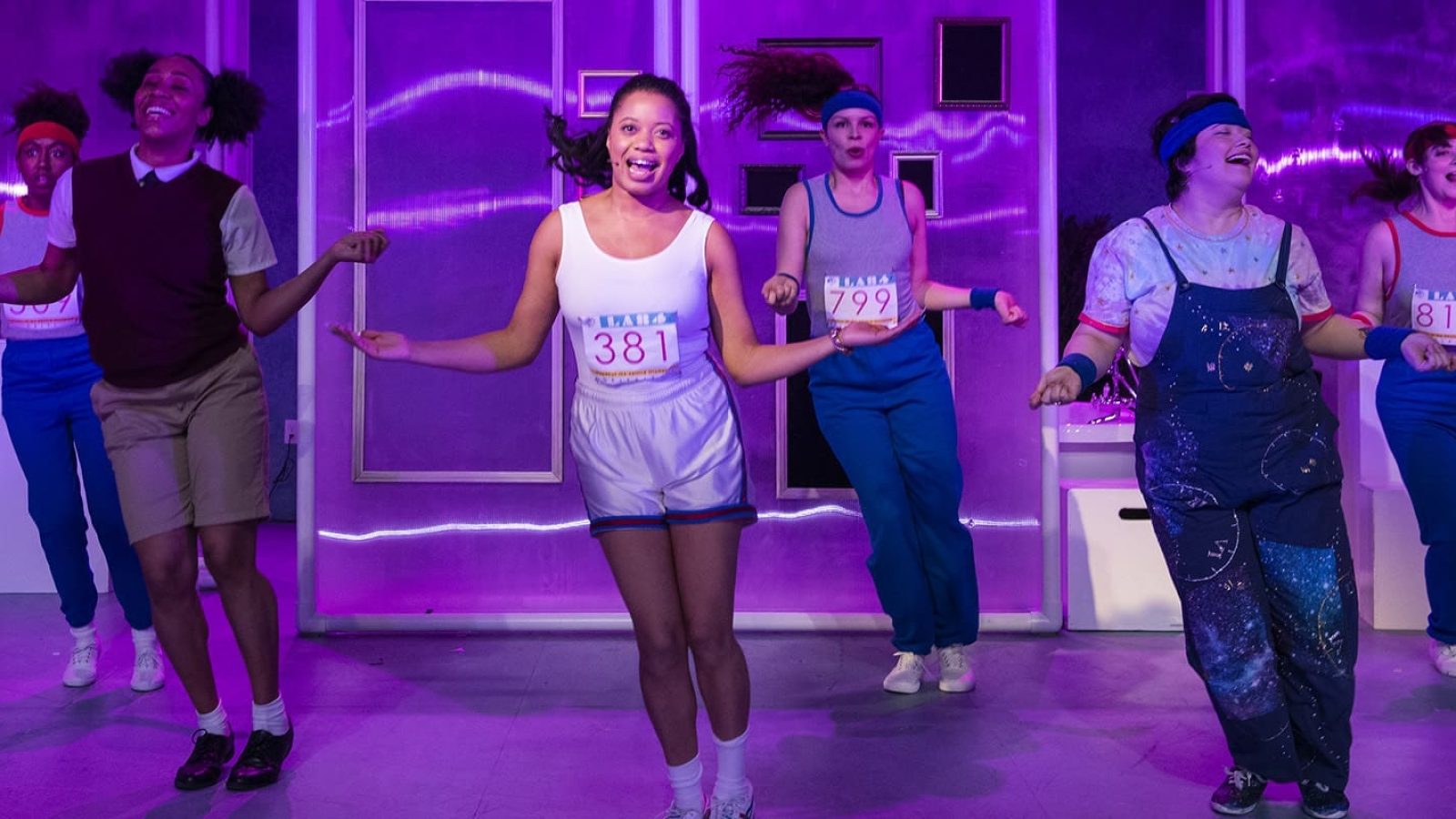 Cast of "She Persisted, The Musical" in vibrant athletic wear, performing a dance number. Numbers 381, 799, and 812 are visible.