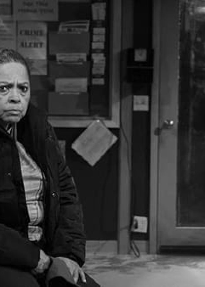 Woman in waiting room with concerned expression, man stands near. Crime Alert sign in background.