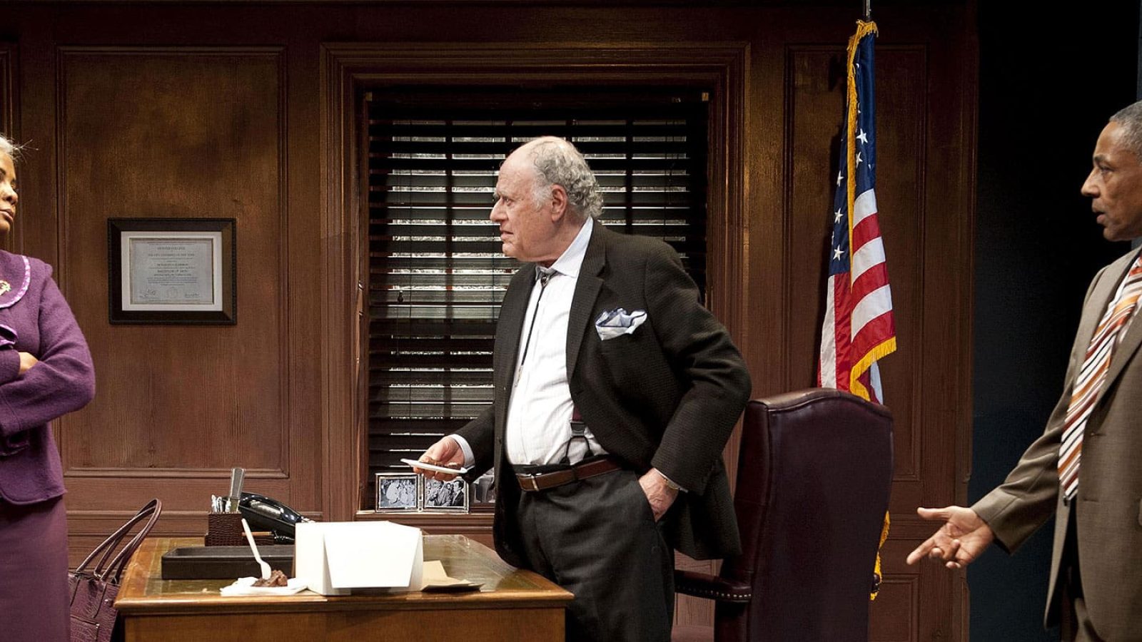 Three people in suits discuss business in an office; one woman, two men.