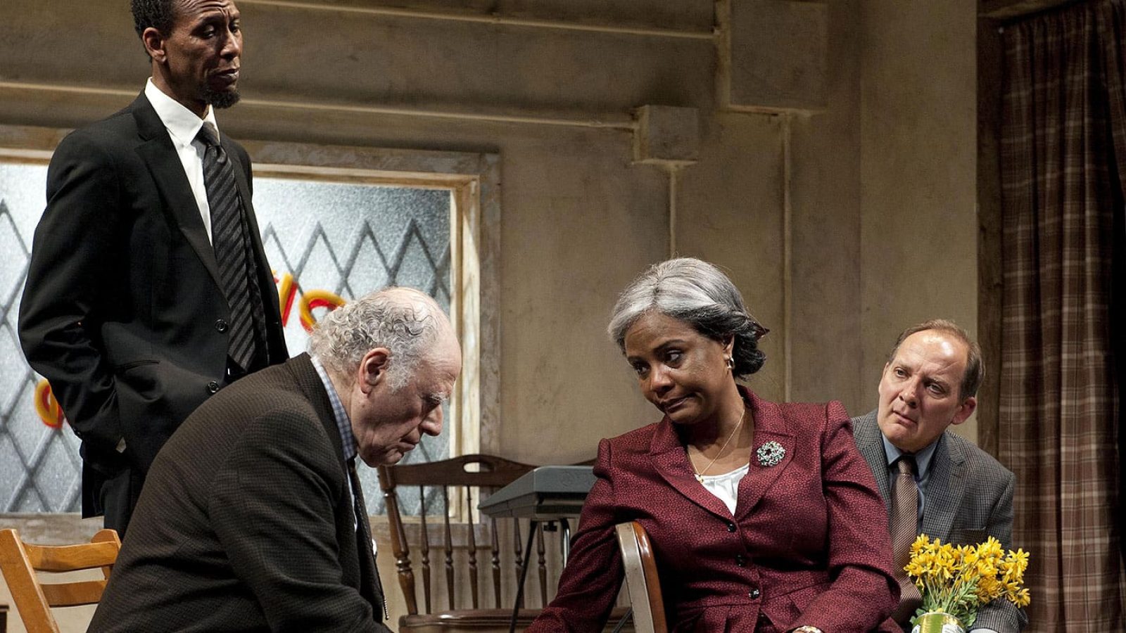 Four adults sit somberly in a storefront church setting; one man stands.