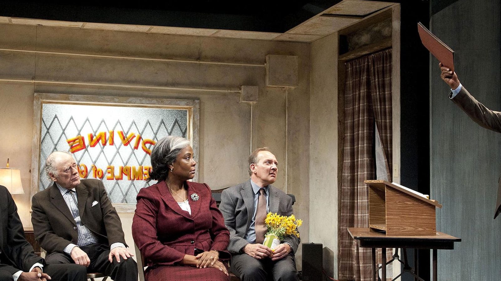 A group of people sit in a storefront church, listening to a man who holds up a file.