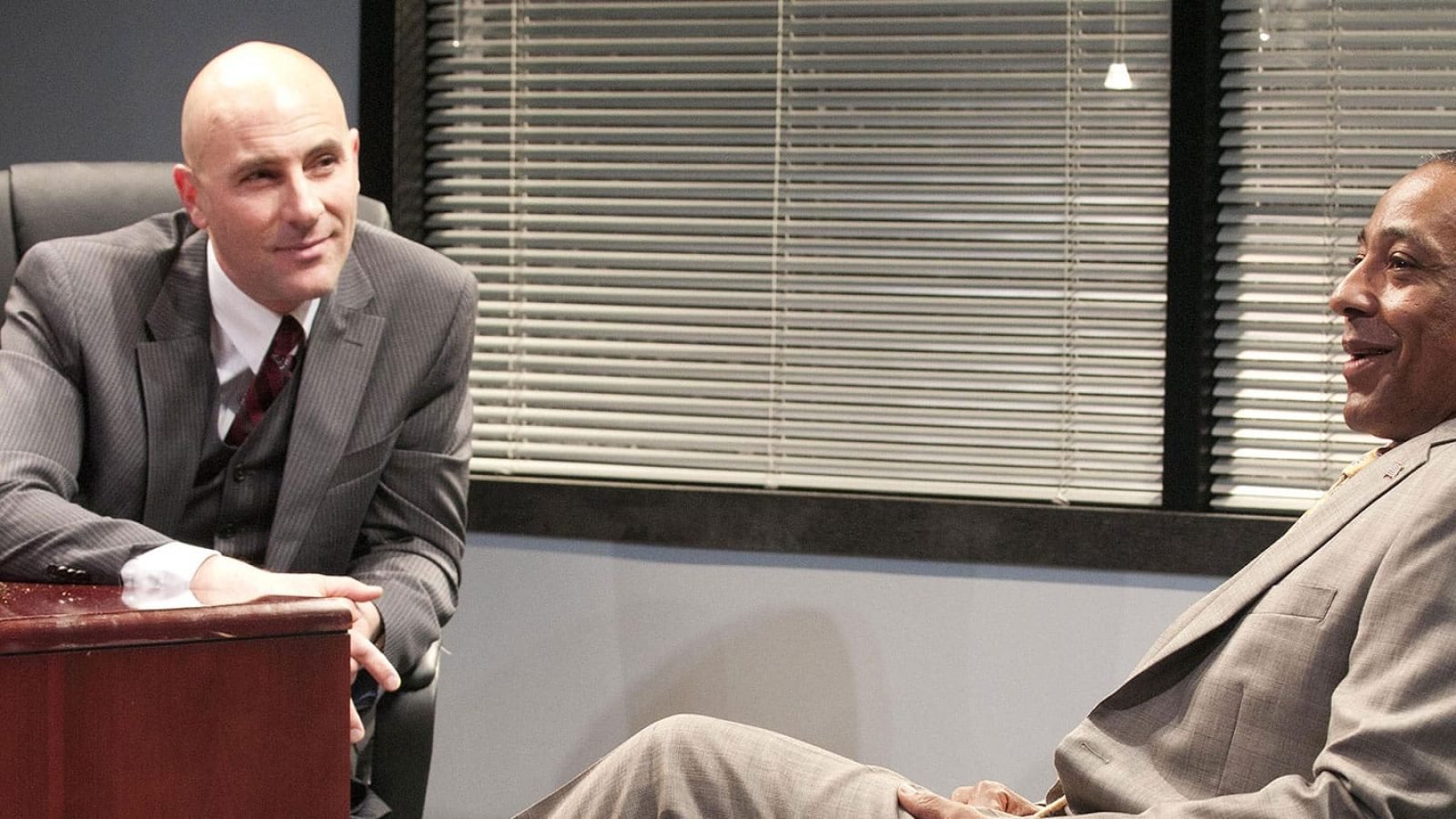 Two men in suits sit across a desk in an office, one smiling.