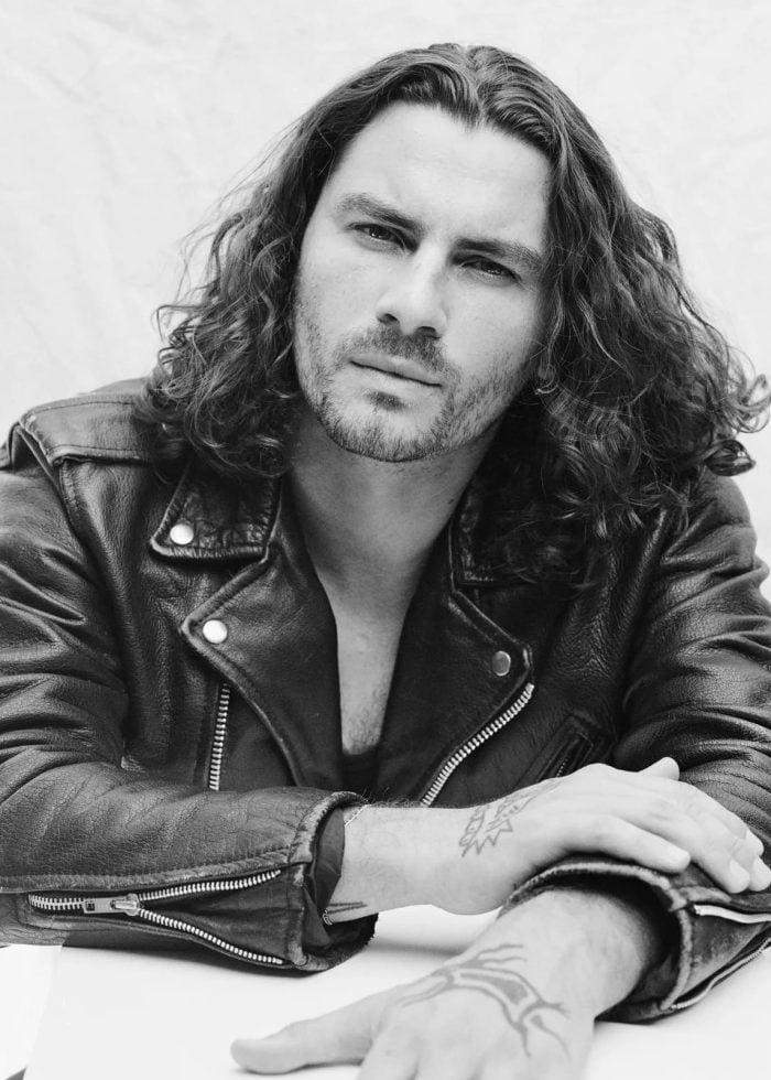 Man with long hair in a leather jacket, looking at the camera. Tattoos are visible on his arm.