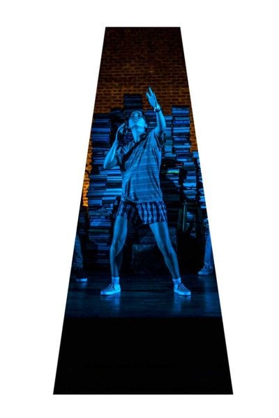 A young person performs on stage, illuminated in blue light, against a backdrop of stacked books.