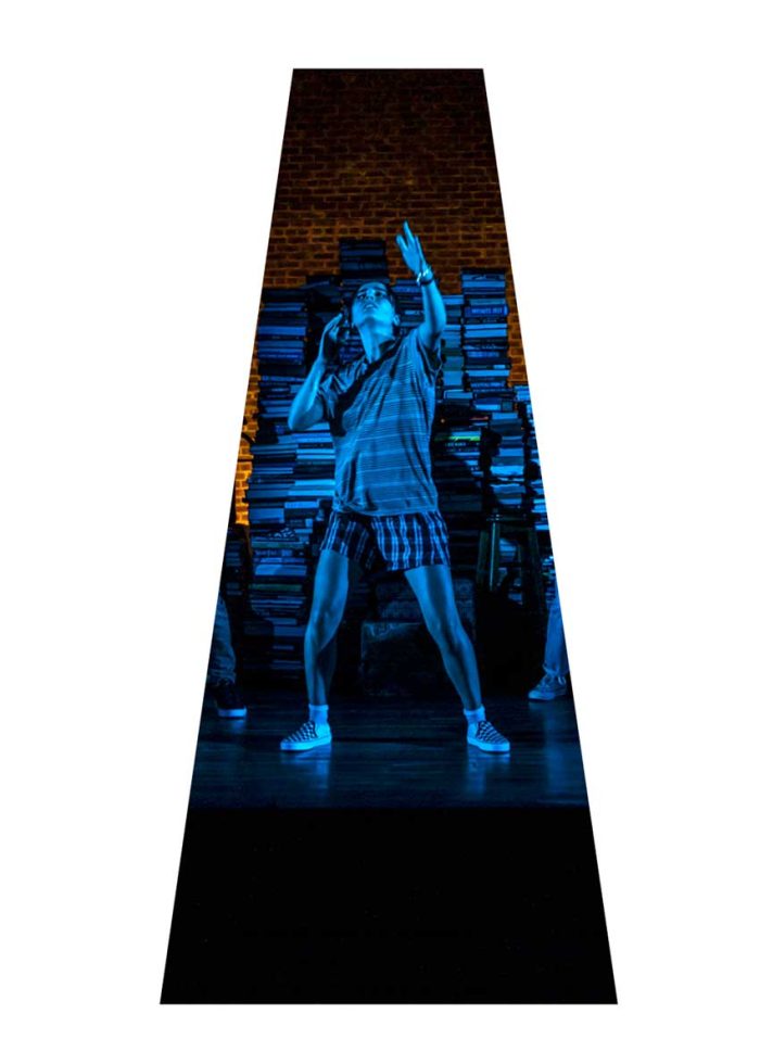A young person performs on stage, illuminated in blue light, against a backdrop of stacked books.