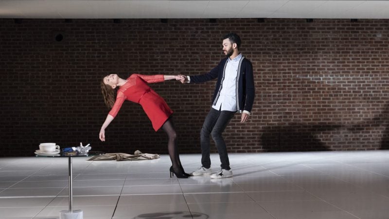 Theater performance: Woman in red dress leans back, held by man in casual attire against a brick wall.