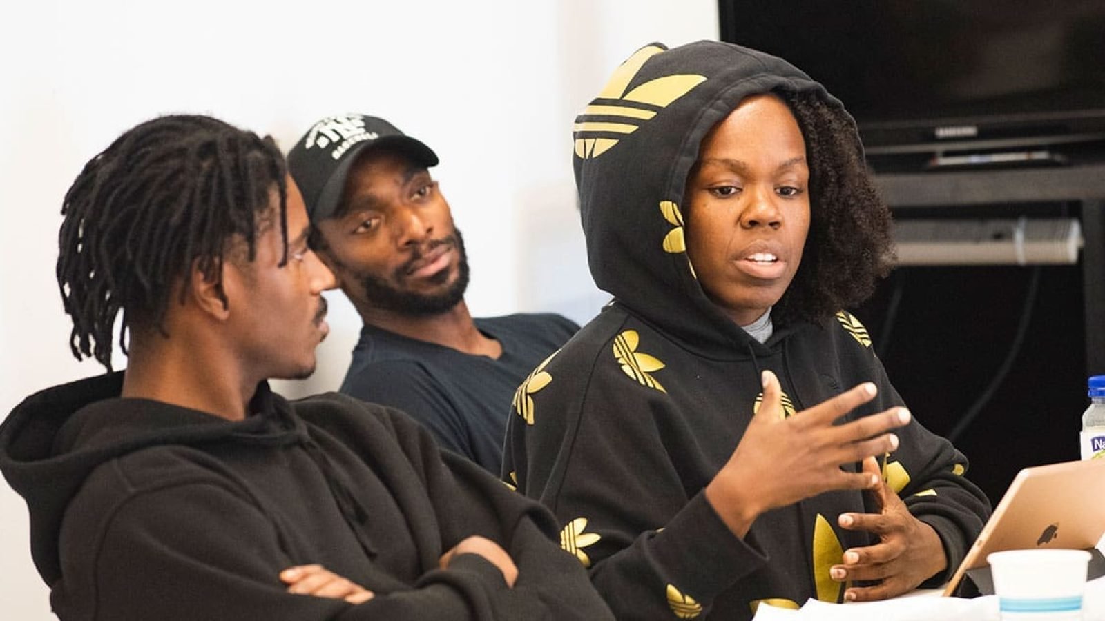 Three young adults in hoodies discuss ideas around a table with a laptop.