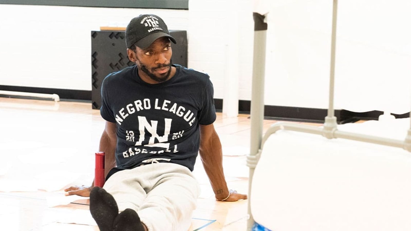Man in Negro League Baseball shirt sits on floor, thoughtful expression.