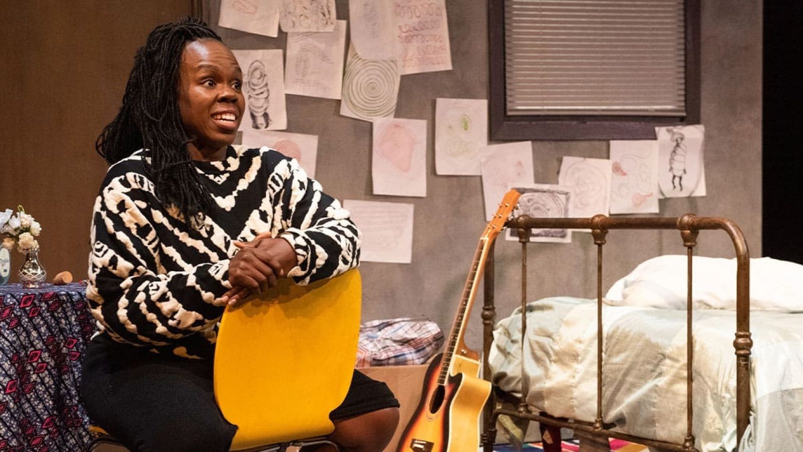 A Black woman sits, looking thoughtfully, in a sparsely furnished room with a bed and guitar.