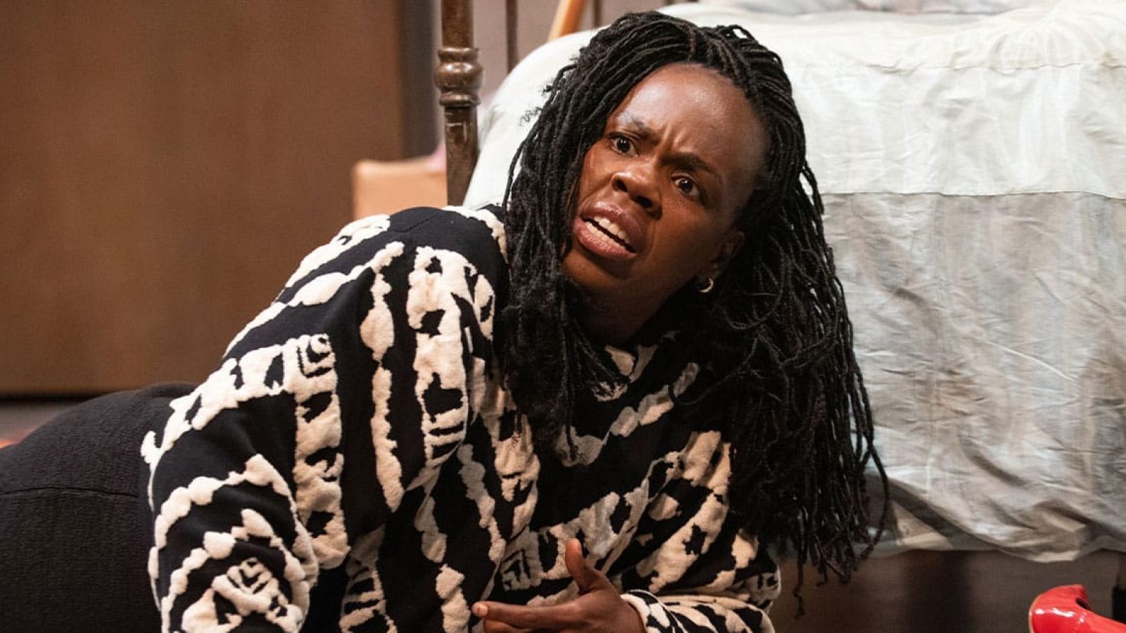 A distraught woman with long braids lies on the floor, looking up in distress.