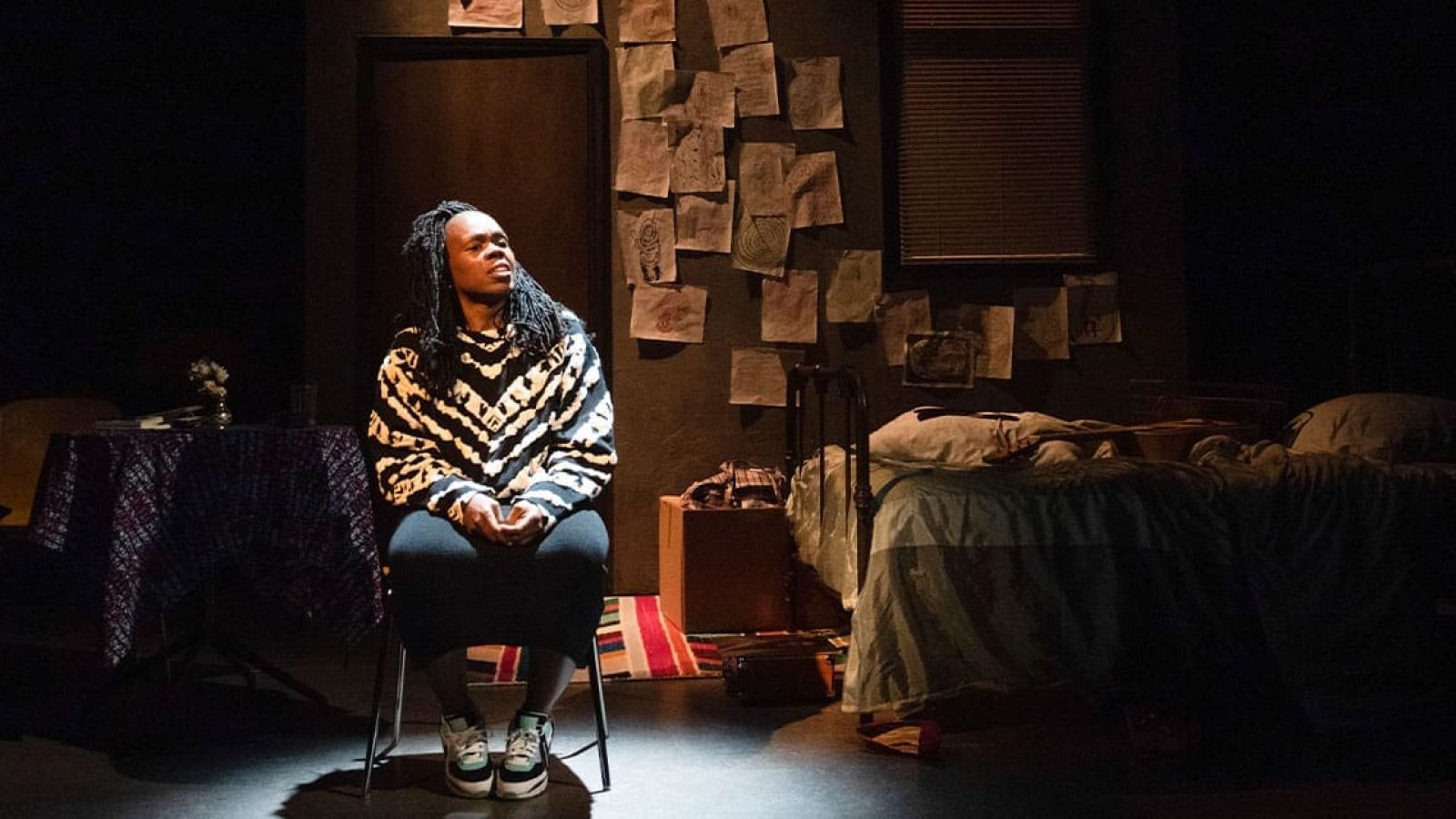 A woman sits pensively in a dimly lit room, surrounded by a bed and personal belongings. The mood is reflective.