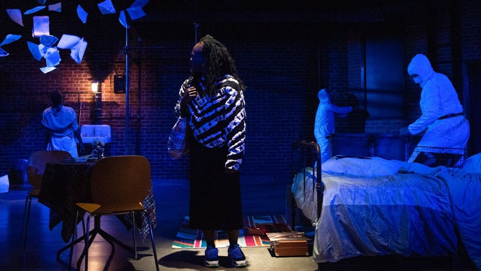 A woman in a black and white sweater stands in a dimly lit room, papers falling around her, as figures in hazmat suits move near a bed. The Last of the Love Letters.