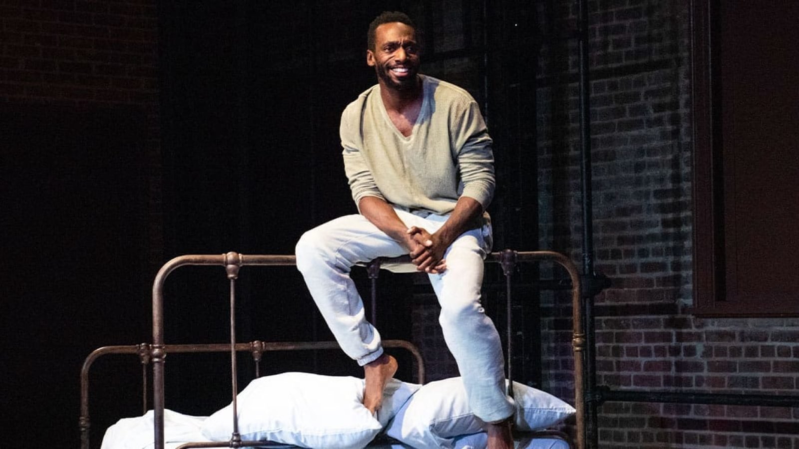 Man sits barefoot on an antique bed frame, smiling.