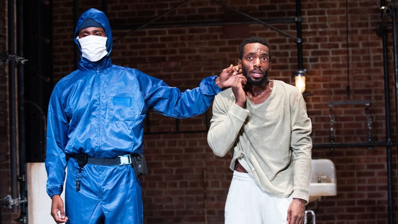 Two men in a stark setting; one in a blue protective suit, the other in distressed clothing, appearing apprehensive.