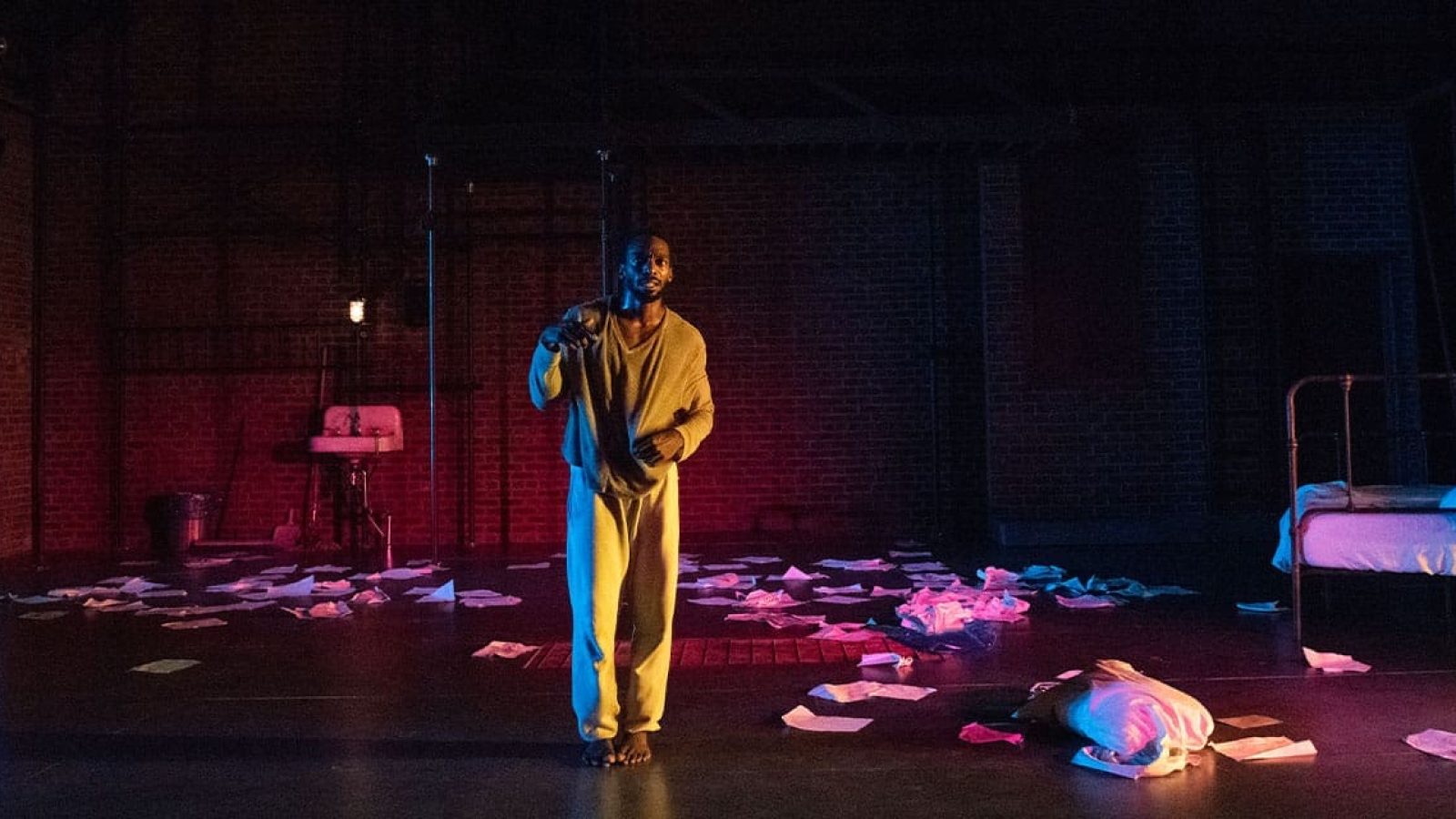 A barefoot man in a loose beige outfit stands on a stage strewn with scattered papers, near a bed. The setting suggests a prison cell or asylum.