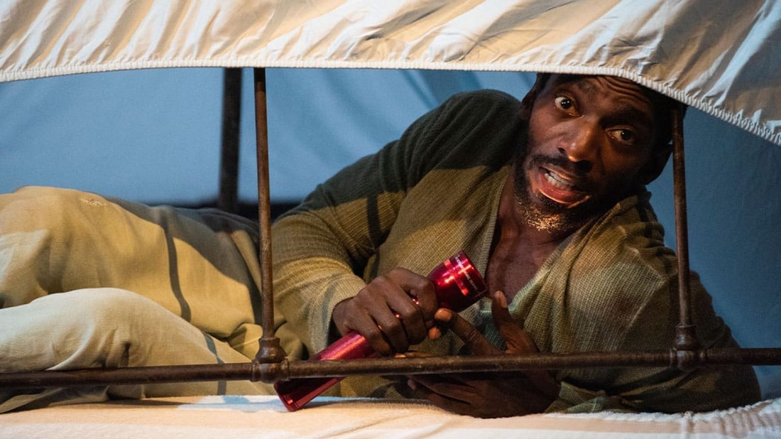 A frightened man hides under a bedsheet, holding a flashlight.