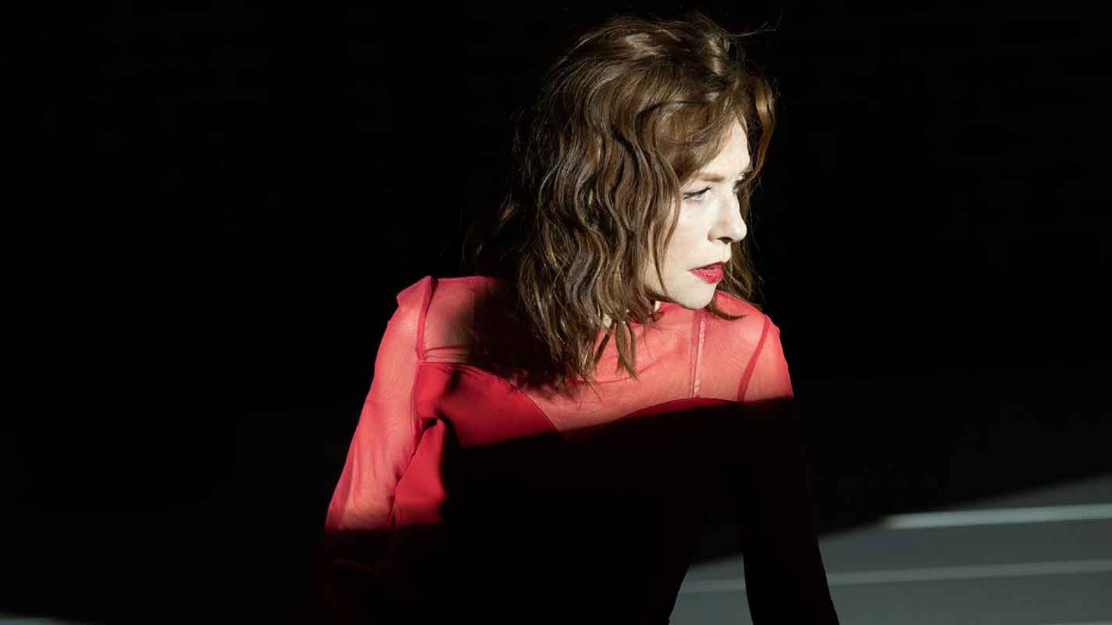 A woman in a red sheer top, looking thoughtfully to her side.