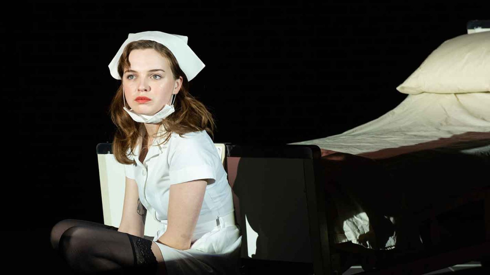 A young nurse sits near an empty hospital bed, looking pensive.
