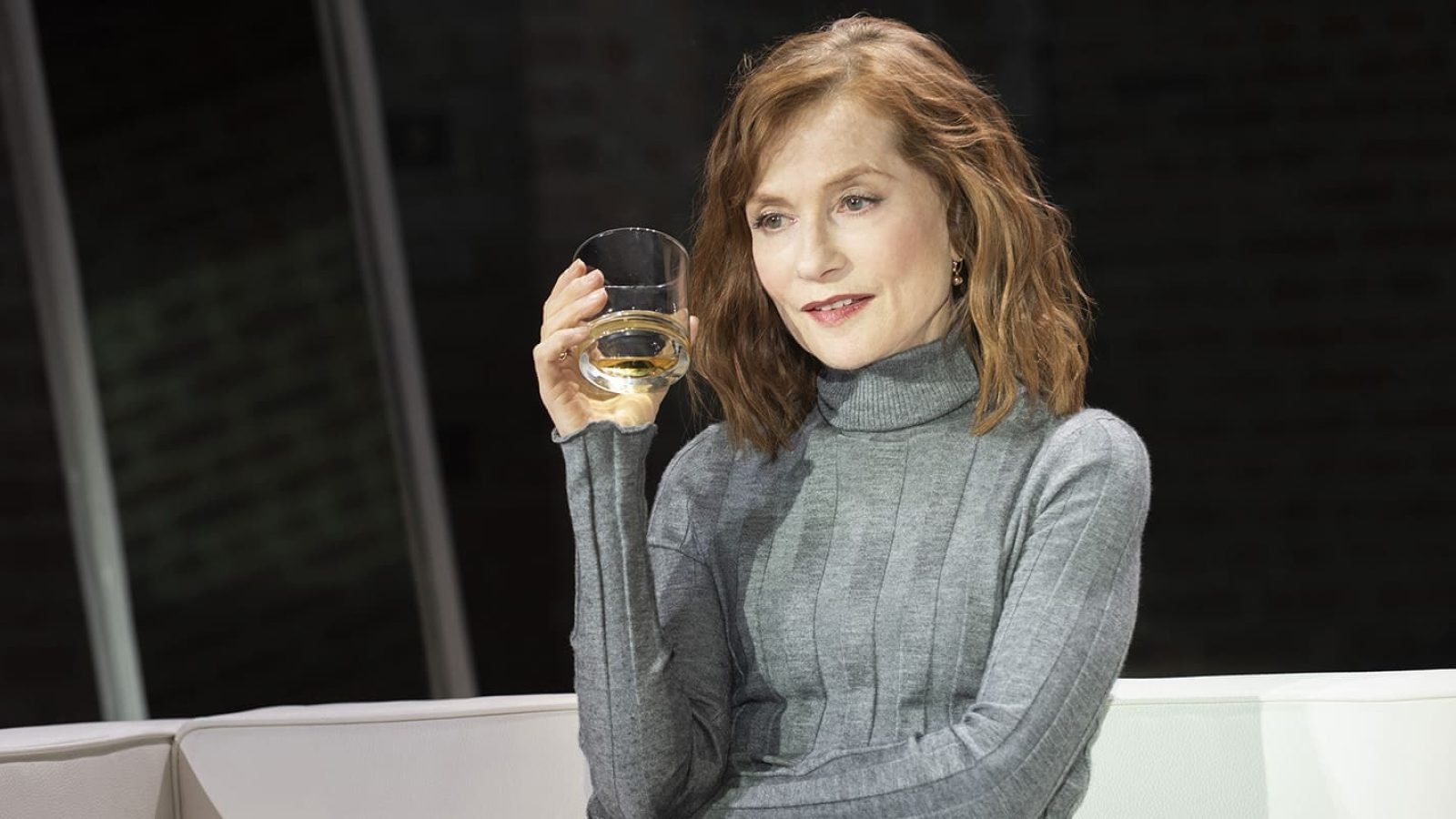 A woman, possibly a mother, sits on a white couch, thoughtfully holding a glass of whiskey.