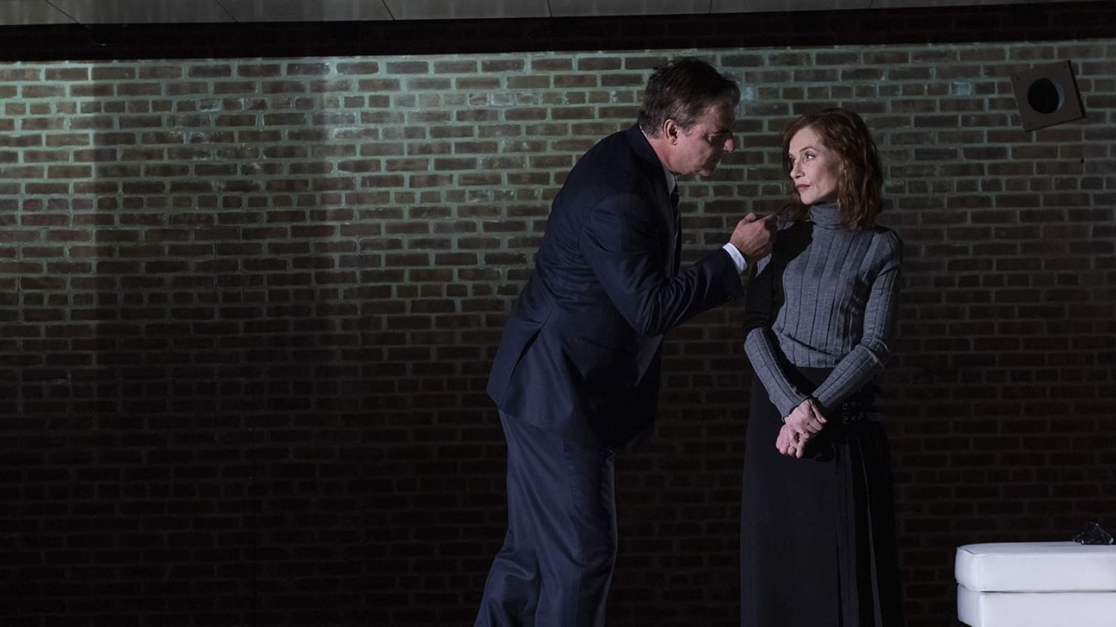 A man and woman in formal attire converse against a brick wall. The woman appears concerned.