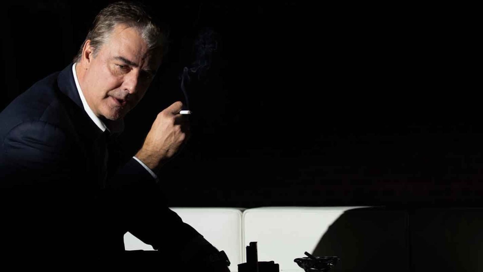 Man in suit smoking a cigarette, sitting on a couch.