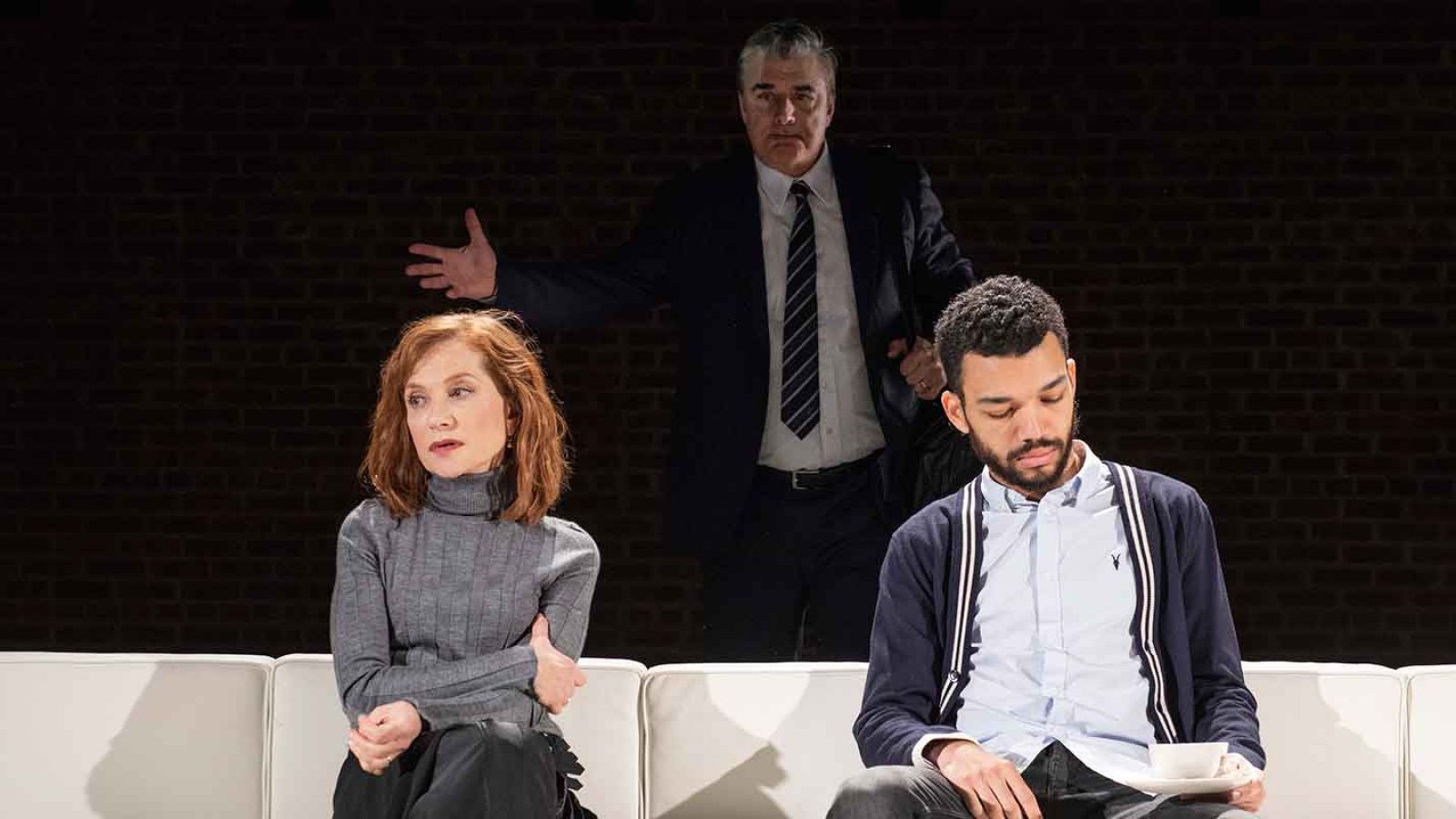 A woman and man sit on a white couch as a man in a suit stands behind them. The Mother.