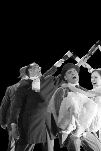 A joyous scene from The Threepenny Opera, featuring actors playfully holding wine bottles above a bride and groom.