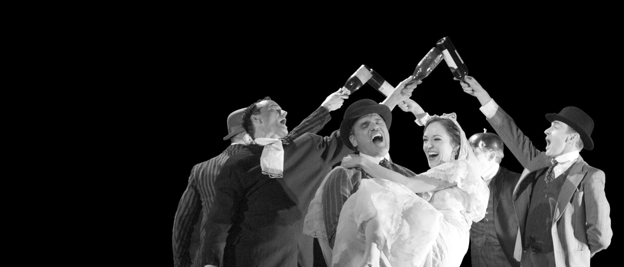 A joyous scene from The Threepenny Opera, featuring actors playfully holding wine bottles above a bride and groom.