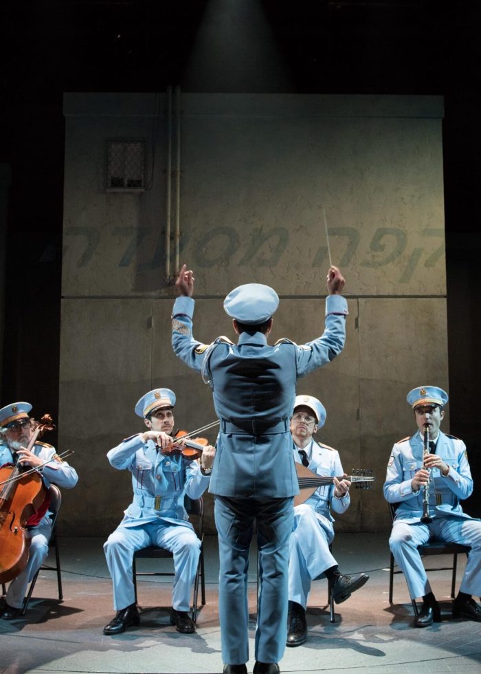 A conductor leads a small orchestra in light blue uniforms, Peter Scanavino-inspired.