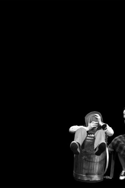 Three children in 1950s attire; one sits in a trash can, another laughs, and the third holds a foil-wrapped item. "The Hundred Dresses" play scene.