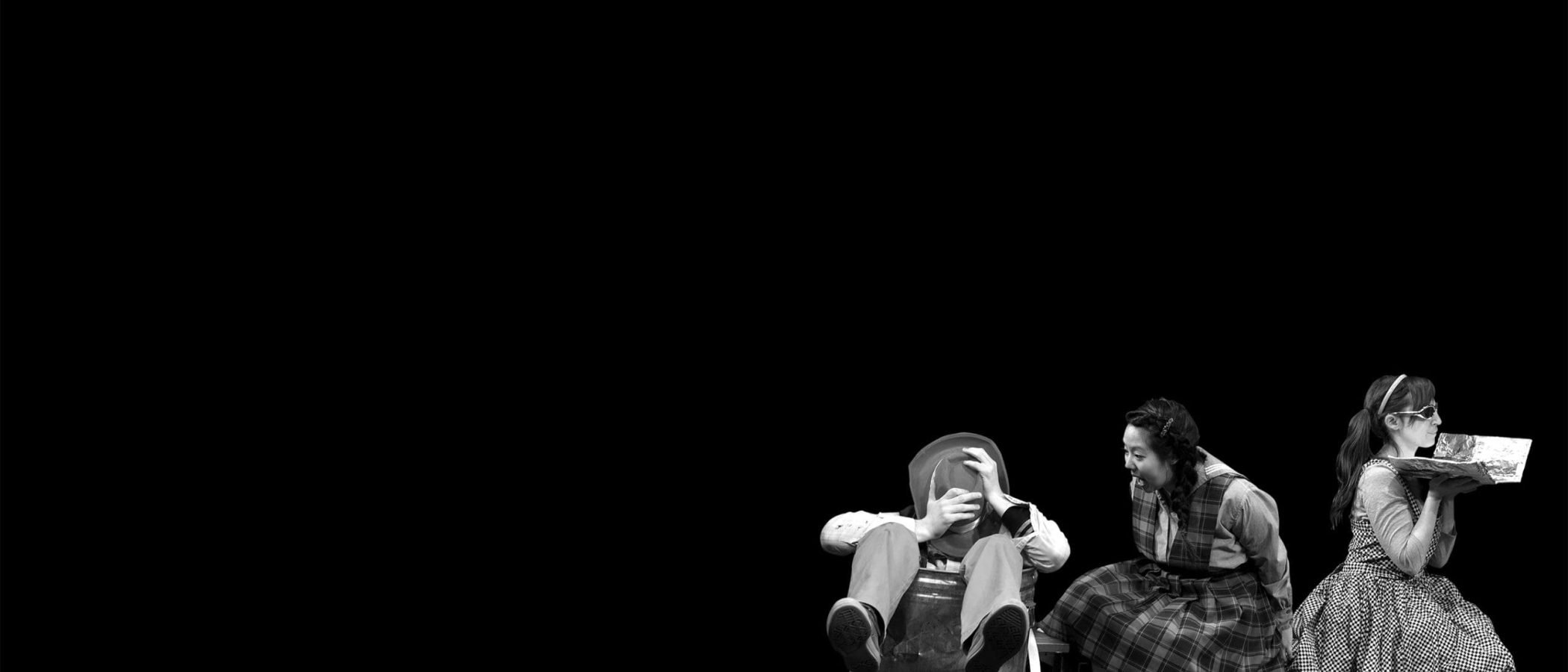 Three children in 1950s attire; one sits in a trash can, another laughs, and the third holds a foil-wrapped item. "The Hundred Dresses" play scene.