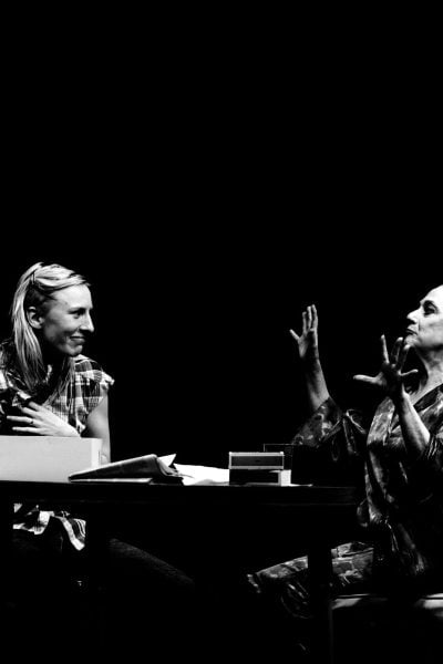 Two women sit at a table, engaged in a serious conversation. One woman appears distraught, while the other gestures emphatically. The Lying Lesson.