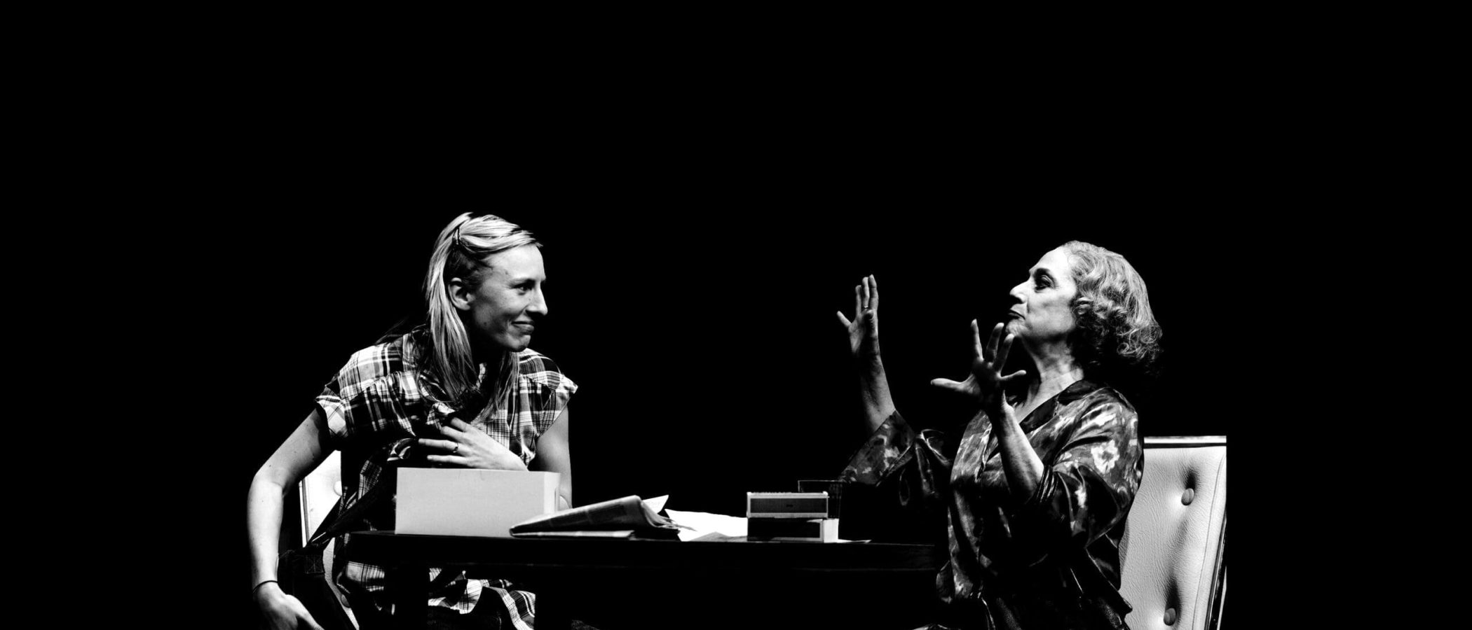 Two women sit at a table, engaged in a serious conversation. One woman appears distraught, while the other gestures emphatically. The Lying Lesson.