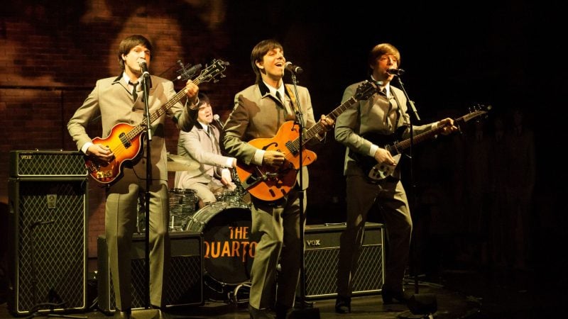 The Quartets band performing on stage, playing guitars and singing. The band is dressed in suits.