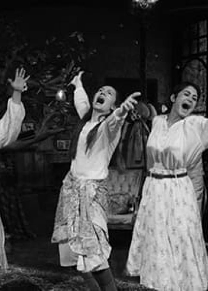 Three women performing on stage, arms raised, singing. Black and white photo.