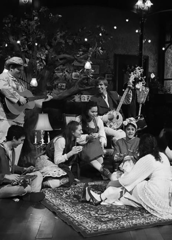 Cast of "This Girl Laughs, Cries – and Grows Up" in a cozy, dimly lit scene, playing music and interacting.
