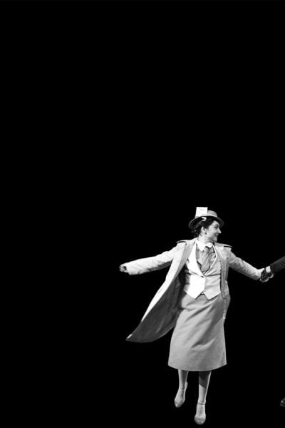A man and woman in 1930s attire playfully dance, holding hands.