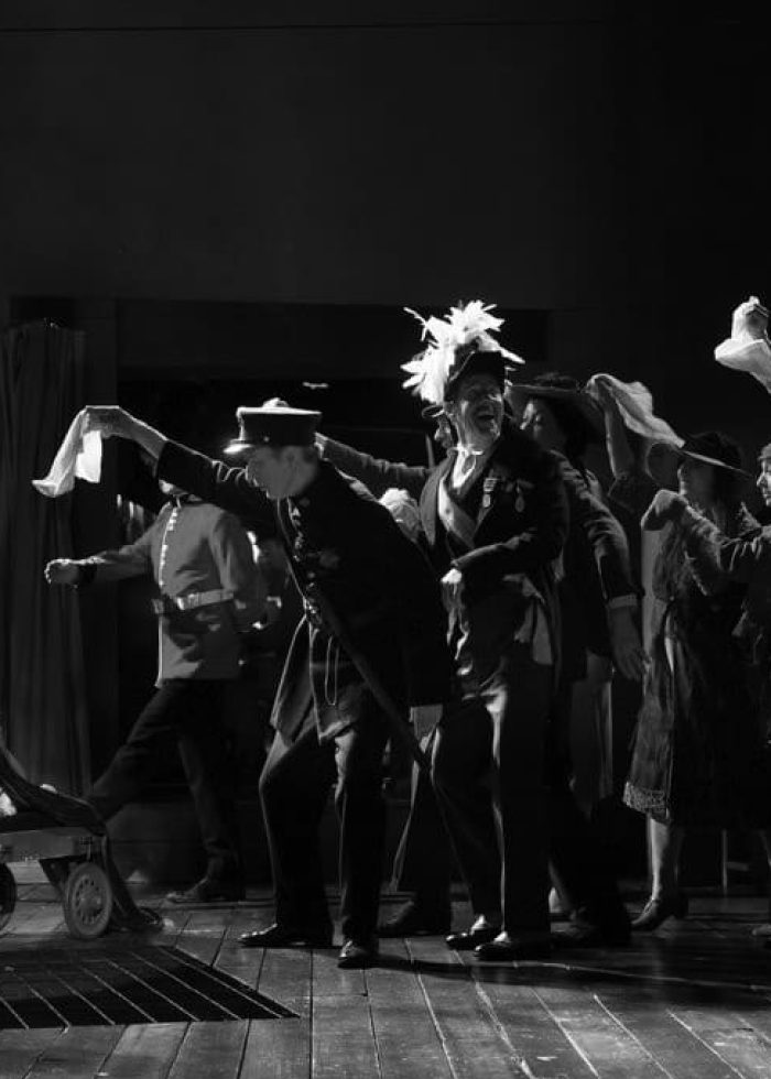 A bulldog wearing a crown on a cart is the center of attention in a theatrical scene. Dogs and Scoundrels, Well Dressed.